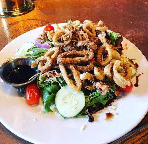 A plate of crispy calamari on a bed of mixed greens, garnished with cherry tomatoes and cucumber slices, served with dipping sauce.