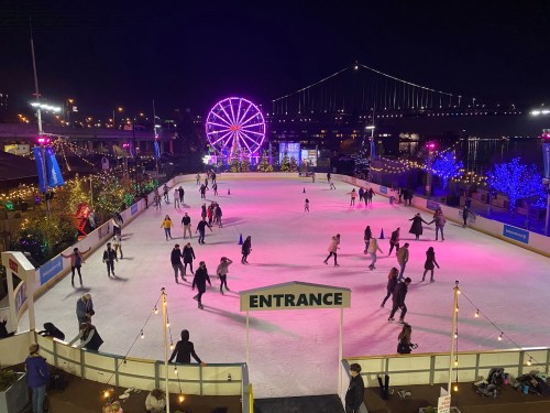 A vibrant outdoor ice skating rink at night, with skaters, colorful lights, and a Ferris wheel in the background.