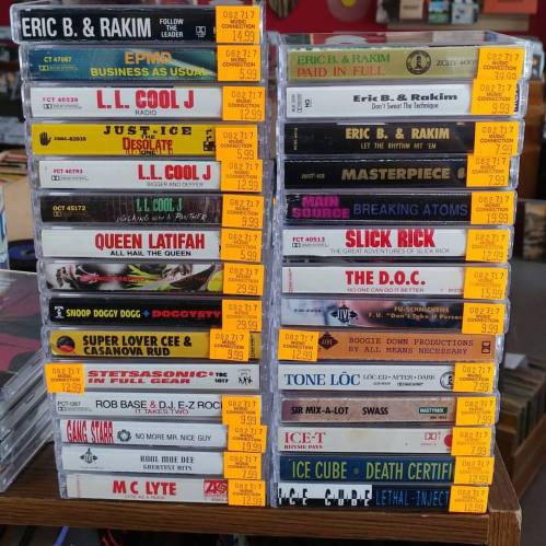 A stack of CDs and cassettes featuring artists like Eric B. & Rakim, LL Cool J, and Ice Cube, displayed in a store.