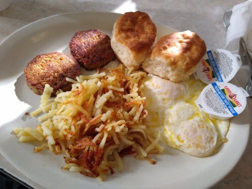 A plate of breakfast featuring two biscuits, hash browns, two eggs, and sausage patties with condiments on the side.