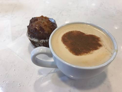 A cup of coffee with a heart-shaped cinnamon design, next to a muffin on a white table.