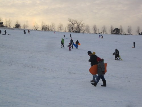People sledding down a snowy hill, with children and adults enjoying the winter day. Trees and a cloudy sky in the background.