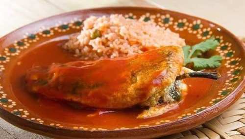 A plate of stuffed chili pepper in red sauce, served with rice and a sprig of cilantro on a decorative dish.