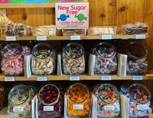 A display of various sugar-free candies in jars on a wooden shelf, with a sign announcing their availability.