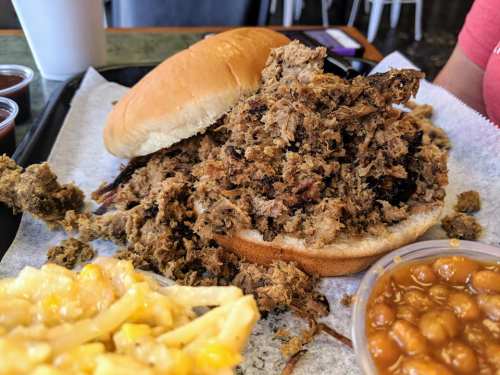 A piled-high pulled pork sandwich on a bun, accompanied by macaroni and cheese and baked beans on a tray.