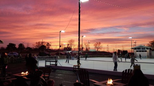 A vibrant sunset over an ice skating rink, with people skating and cozy fire pits in the foreground.
