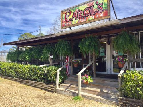 A rustic market with a sign reading "Carol's Marketplace," featuring ferns and flowers on the porch and a welcoming entrance.