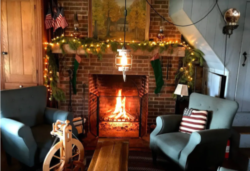 Cozy living room with a fireplace, decorated with stockings, greenery, and string lights, featuring two armchairs and a wooden table.
