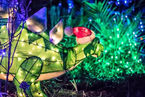 A festive dinosaur decoration wearing a Santa hat, surrounded by colorful holiday lights and greenery.