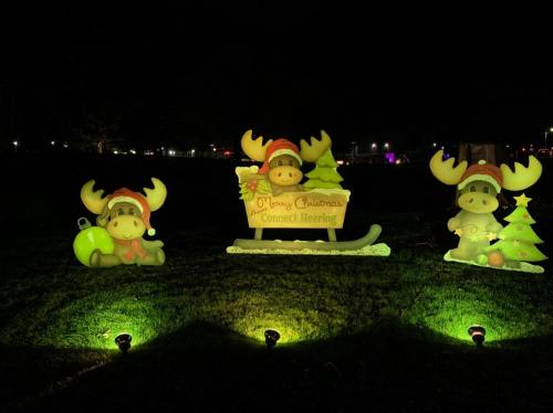 Three festive moose decorations with Christmas hats, a sign reading "Merry Christmas," and a small tree, illuminated at night.
