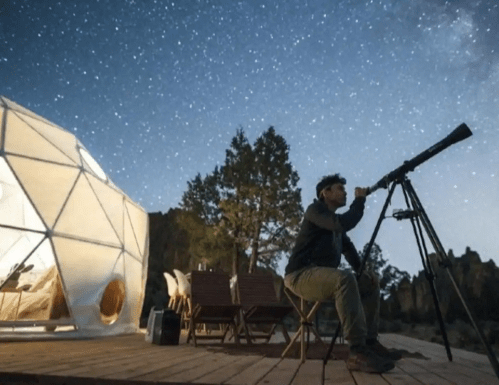 A person observes the night sky through a telescope near a geodesic dome under a starry sky.