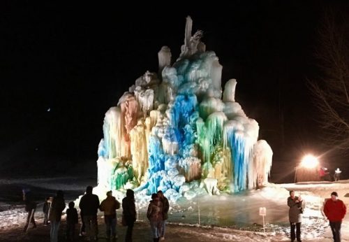 A colorful ice formation illuminated at night, with people gathered around it, creating a festive winter atmosphere.