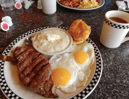 A hearty breakfast plate with steak, two fried eggs, grits with butter, a biscuit, and a cup of coffee.