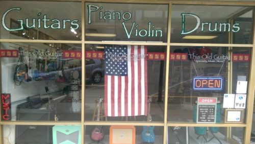 Storefront of a music shop featuring instruments like guitars, piano, and drums, with an American flag and "OPEN" sign.