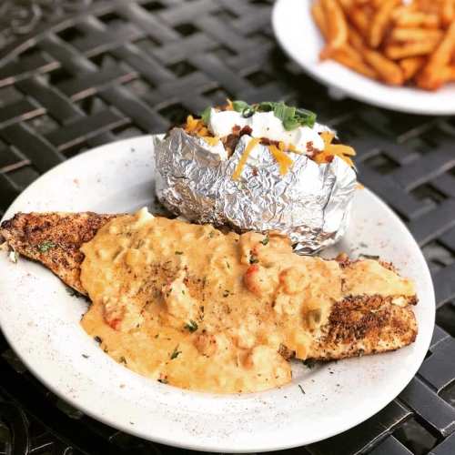 A seasoned fish fillet topped with creamy sauce, served with a baked potato and a side of sweet potato fries.