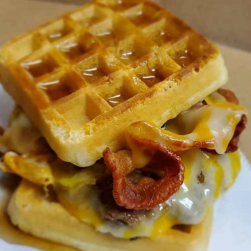 A stacked waffle sandwich with melted cheese, crispy bacon, and a savory filling, drizzled with syrup.