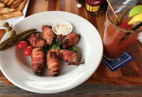 A plate of bacon-wrapped jalapeño poppers with dipping sauce, cherry tomatoes, and pickles, alongside a cocktail.