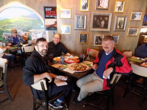 Three men sit at a table in a restaurant, enjoying a meal surrounded by framed photos on the wall.