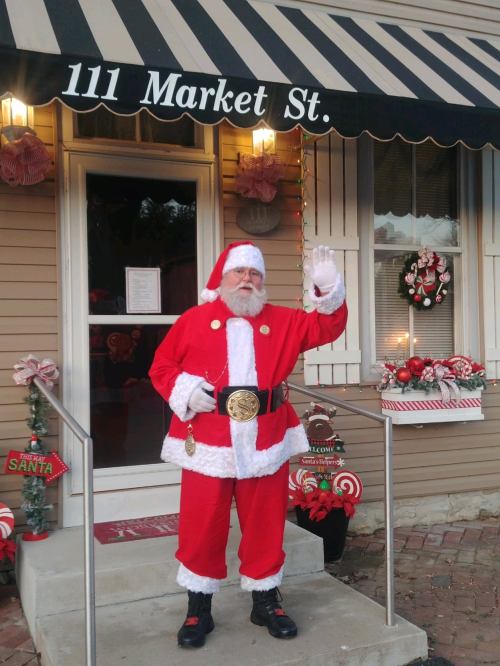Santa Claus waves outside a festive storefront decorated for the holidays, wearing a red suit and black belt.