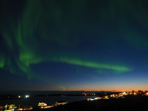 A vibrant display of green auroras dances across a dark sky above a coastal town at dusk.
