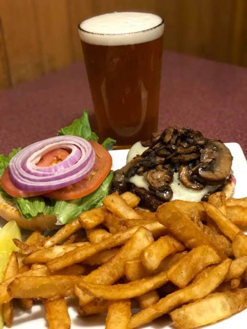 A plate with a burger topped with mushrooms, lettuce, tomato, and onion, served with fries and a glass of beer.