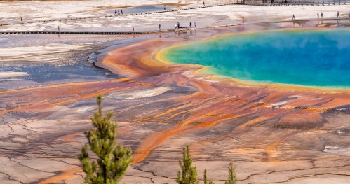 A vibrant geothermal pool with blue and orange hues, surrounded by a barren landscape and visitors in the distance.