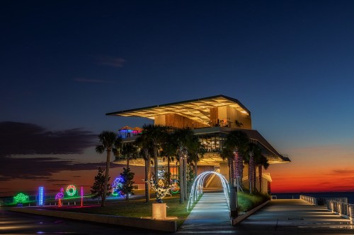 A modern building illuminated with colorful lights at dusk, surrounded by palm trees and a vibrant sunset sky.