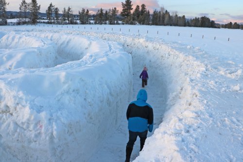 A child and an adult walk through a snowy maze in a winter landscape, surrounded by trees and a clear sky.