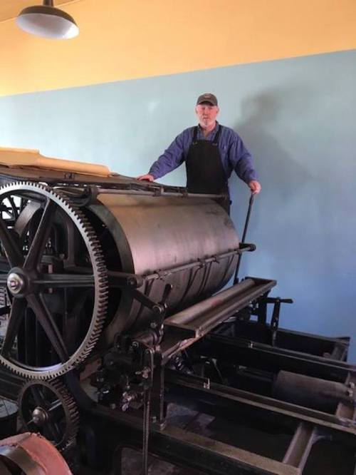 A man stands on a vintage printing press, operating it in a workshop with blue walls.