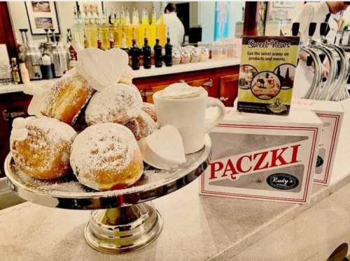 A silver platter piled with powdered sugar-dusted paczki, alongside a cup and a promotional sign for a bakery.