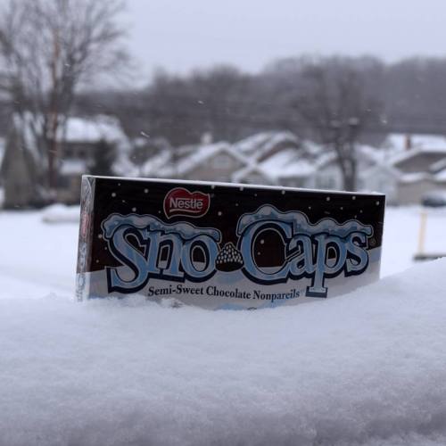 A box of Nestlé Sno Caps chocolate nonpareils resting on a snowy surface with houses in the background.