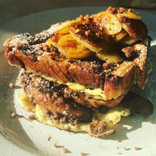 A stack of French toast topped with sliced bananas, nuts, and a dusting of powdered sugar on a white plate.