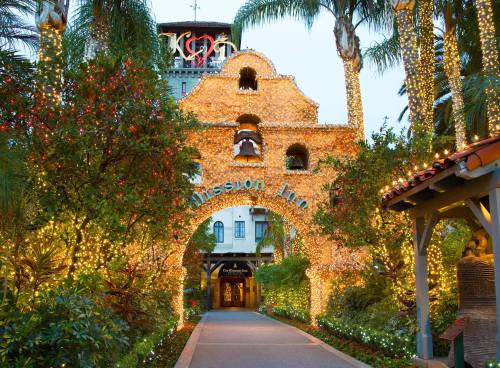 A beautifully lit archway adorned with holiday lights, leading to the Mission Inn, surrounded by palm trees and greenery.