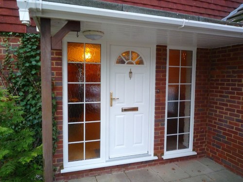A white front door with glass panels, flanked by two windows, set against a brick wall and covered porch.
