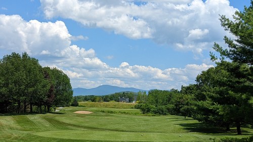 A scenic view of a golf course with lush green grass, trees, and distant mountains under a partly cloudy sky.