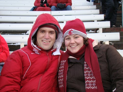 A smiling couple in winter attire, sitting together in a stadium with empty bleachers behind them.