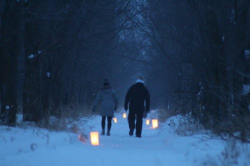 A couple walks hand in hand down a snowy path lined with glowing lanterns, surrounded by dark trees.