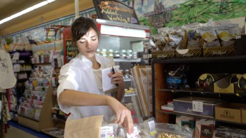 A woman in a white chef's coat organizes snacks in a store filled with various products and displays.