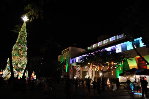 A festive scene at night featuring a large decorated Christmas tree and a building adorned with colorful lights.