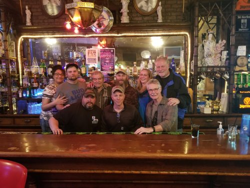 A group of people poses together at a bar, smiling, with colorful bottles and lights in the background.