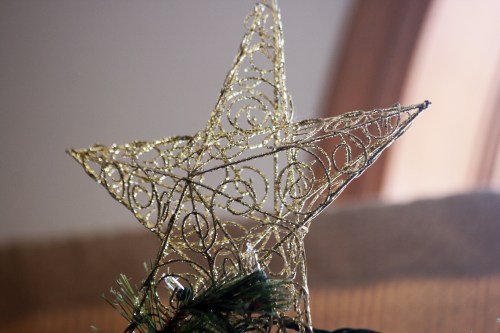 A decorative gold star with intricate swirls atop a Christmas tree, set against a softly lit background.