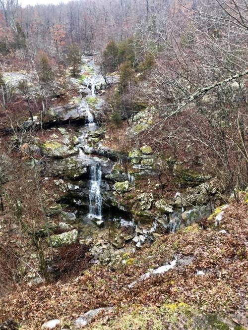 A serene waterfall cascading over rocky terrain, surrounded by bare trees and autumn foliage in a forested area.