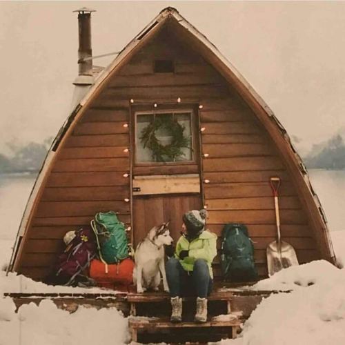 A cozy wooden cabin in the snow, with a person sitting beside a dog and colorful backpacks nearby.