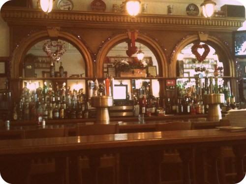 A cozy bar with a wooden counter, shelves lined with various bottles, and decorative hearts on the mirrors.