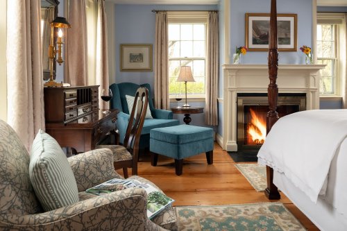 Cozy bedroom with blue walls, a fireplace, armchairs, and a wooden desk, featuring warm lighting and floral decor.
