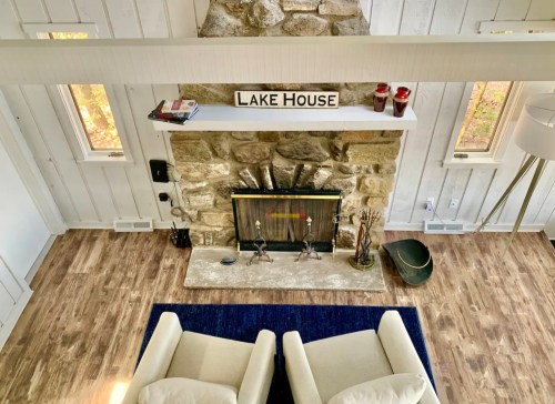 Cozy living room with a stone fireplace, wooden floors, and a "Lake House" sign on the mantel. Two chairs in view.