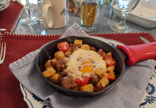 A cast iron skillet filled with potatoes, sausage, cherry tomatoes, and a sunny-side-up egg on a blue napkin.
