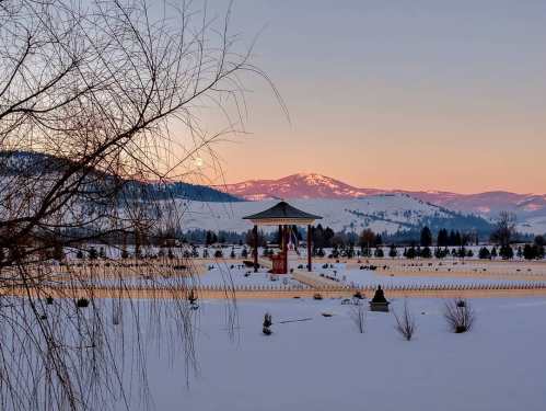 A serene winter landscape with a gazebo, snow-covered ground, and mountains at sunset, creating a peaceful atmosphere.