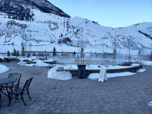 A snowy landscape with mountains in the background, featuring a steaming hot spring and a stone patio with chairs.