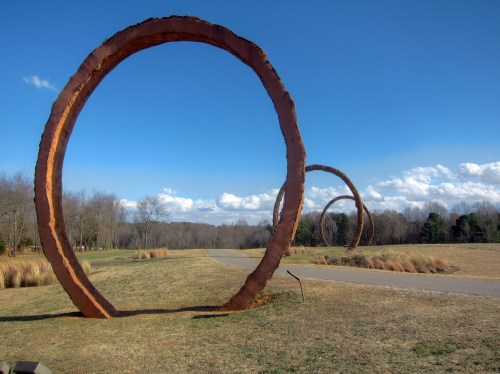 Three large circular sculptures made of wood stand in a grassy field under a clear blue sky with scattered clouds.